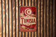 A vintage-style metal sign with the word "TUNISIA" prominently displayed, featuring a crescent moon and star symbol, and a decorative border with a desert landscape and buildings.