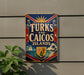 A vintage metal sign with the text "TURKS AND CAICOS ISLANDS" displayed on a wall, with a sailboat and palm trees in the background.
