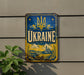 a vintage-style metal sign with the word "UKRAINE" prominently displayed, along with a decorative border and a depiction of a castle. The sign is mounted on a wall, and there is a plant visible to the left of the sign.