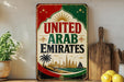 A vintage-style sign that reads "UNITED ARAB EMIRATES" with a colorful design featuring a sun, stars, and a palm tree. The sign is displayed on a shelf next to a wooden cutting board and a potted plant.