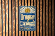 A vintage-style metal sign with the word "Uruguay" prominently displayed, featuring a sun and palm trees, against a wooden background.