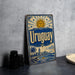 A vintage-style metal sign with the word "Uruguay" prominently displayed, featuring a sun and palm trees, is leaning against a concrete wall.