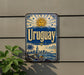 a vintage-style metal sign with the word "Uruguay" prominently displayed, along with a sun and palm tree illustration. The sign is mounted on a wall, and there is a plant visible to the left of the sign.