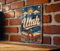A vintage-style metal sign with the word "Utah" prominently displayed, featuring a mountain landscape and sun-like symbols.
