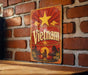 A vintage-style metal sign with the word "Vietnam" prominently displayed, featuring a red and gold color scheme with a yellow star and a sunset-like background.