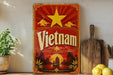 a vintage-style sign with the word "Vietnam" prominently displayed, along with a sun and clouds design. The sign is placed on a shelf next to a wooden cutting board and a potted plant.