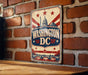 A vintage-style metal sign with the text "WASHINGTON DC" displayed prominently, featuring a brick wall and a bridge in the background.
