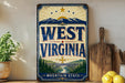 A vintage-style metal sign with the text "WEST VIRGINIA" and a scenic illustration of mountains, trees, and a bridge, displayed on a shelf next to a wooden cutting board and some potted plants.
