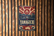 A vintage-style metal sign with the word "YAMAGUCHI" prominently displayed, featuring a red and white design with a sunburst pattern and Japanese-style architecture.