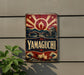 A vintage-style sign with a red and blue design, featuring a sunburst pattern and a Japanese-style illustration, mounted on a wall.