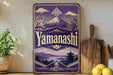 A vintage-style sign for Yamazaki, featuring a mountain landscape with cherry blossoms and a pagoda, is displayed on a shelf next to a wooden cutting board and some lemons.