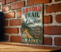 A vintage metal sign for the "Arnold Trail to Quebec" in Maine, featuring a scenic road and wilderness landscape.