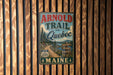 A vintage metal sign for the "Arnold Trail to Quebec" in Maine, featuring a scenic road surrounded by pine trees and mountains.