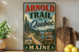 A vintage-style sign for the "Arnold Trail to Quebec" in Maine, featuring a scenic road sign and a mountain landscape.