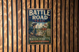 A vintage metal sign with the title "Battle Road Trail" and an illustration of a group of people on a road, mounted on a wooden wall.