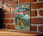 A vintage metal sign for the Bay Circuit Trail in Massachusetts, featuring a scenic illustration of a coastal landscape with a sailboat.
