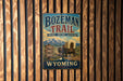 A vintage metal sign for Bozeman Trail, featuring a wagon train and mountains in the background, is displayed on a wooden wall.