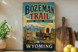 a vintage-style sign for Bozeman Trail, which is a historic trail in Wyoming. The sign has a picture of a covered wagon and mountains in the background, along with directional signs pointing to various locations. The sign is displayed on a shelf next to a wooden cutting board and some lemons.