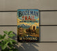 A vintage-style metal sign for Bozeman Trail, featuring a wagon train and mountains in the background, mounted on a wall next to a plant.