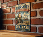 A vintage-style metal sign featuring a portrait of a bearded man with a beard, along with text indicating the location of the Chesapeake Historic Trail in Delaware.
