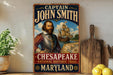 A vintage-style metal sign featuring a portrait of a bearded man, with the text "CAPTAIN JOHN SMITH" and "CHESAPEAKE - NATIONAL HISTORIC TRAIL - MARYLAND" displayed prominently.