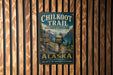 A metal sign on a wooden wall that reads "CHILKOOT TRAIL HISTORIC CORRIOR ALASKA" with a picture of a hiker and mountains in the background.