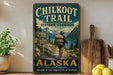 A vintage-style metal sign for the Chilkoot Trail Historic Corridor in Alaska, featuring a hiker on a trail surrounded by mountains and a scenic landscape.