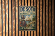 A vintage metal sign for the De Soto National Memorial Trail in Florida, featuring a historical illustration of a man in period attire and a ship in the background.