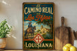 A vintage-style metal sign with the text "El Camino Real de Los Tejas Louisiana" displayed on it, featuring an illustration of a cowboy on a horse and a Spanish-style building.