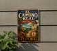 a vintage-style metal sign with a tropical scene, mounted on a wall next to a plant.