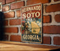 A vintage-style metal sign with the text "Hernando de Soto Trail Georgia" and an illustration of a man in a historical-looking outfit, mounted on a brick wall.