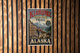 A vintage-style metal sign for Klondike Gold Rush Trail in Alaska, featuring a mountain landscape and a person in a red jacket.