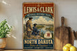 A vintage-style metal sign for the Lewis & Clark National Historic Trail, featuring two figures in historical attire standing on a cliff overlooking a river.