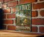 A vintage-style metal sign for the Old Trace Historic Path in Mississippi, featuring a scenic illustration of a path surrounded by trees and a body of water.