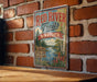 A vintage-style metal sign for the Red River Trail in Minnesota, featuring a scenic image of a river surrounded by trees and flowers.