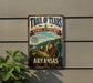 a vintage-style sign for the Trail of Tears National Historic Trail in Arkansas, with a group of people walking along a path through a scenic landscape.
