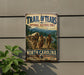 a vintage-style metal sign with a painting of a group of people walking along a trail, set against a backdrop of mountains and a river.
