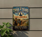a vintage-style sign for the Trail of Tears, which is a National Historic Trail in Oklahoma. The sign depicts a group of people, likely Native Americans, traveling down a dirt road with a horse-drawn wagon. The sign is mounted on a wall, and there is a plant to the left of the sign.