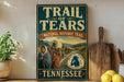 A vintage-style sign for the Trail of Tears, a historic event in the United States, featuring a painting of people in traditional clothing and a wagon. The sign is displayed on a shelf next to a wooden cutting board and some lemons.