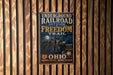 a vintage-style metal sign with the text "Underground Railroad Network to Freedom Trail" and "OHIO" printed on it, hanging on a wooden wall.
