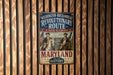 A vintage-style metal sign with the text "WASHINGTON-ROCCHAMBEAU REVOLUTIONARY ROUTE A PATH TO LIBERTY MARYLAND" and an illustration of two men on horseback, mounted on a wooden wall.
