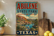 A vintage-style sign for Abilene State Park in Texas, featuring a scenic landscape with mountains, cacti, and a river.