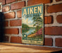 A vintage-style metal sign for Aiken State Park, featuring a scenic landscape with a lake, trees, and a palm tree.