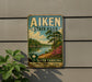 A vintage-style sign for Aiken State Park, featuring a scenic landscape with a river, trees, and a palm tree.