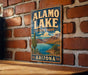 A vintage-style metal sign for Alamo Lake State Park in Arizona, featuring a desert landscape with a cactus and a lake.