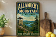 A vintage-style metal sign for Allamuchy Mountain State Park, featuring a scenic mountain landscape with a lookout tower, is displayed on a shelf next to a wooden cutting board and some potted plants.