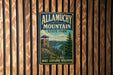 A vintage metal sign for Allamuchy Mountain State Park, featuring a scenic mountain landscape with a lookout tower, is displayed on a wooden wall.