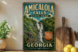 A vintage-style sign for Amicola Falls State Park in Georgia, featuring a waterfall and lush greenery, is displayed on a shelf next to a wooden cutting board and some lemons.