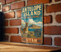A vintage metal sign for Antelope Island State Park in Utah, featuring an illustration of a mountain range and a gazelle.