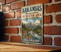A vintage metal sign for Arkansas Headwaters Recreation Area in Colorado, featuring a scenic mountain landscape with a river and pine trees.